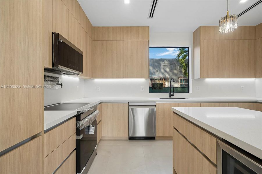 Kitchen featuring flat-panel wood-finish cabinetry, white solid-surface countertops, an integrated center island, stainless steel appliances, and under-cabinet lighting Kitchen featuring flat-panel wood-finish cabinetry, white solid-surface countertops, an integrated center island, stainless steel appliances, and under-cabinet lighting