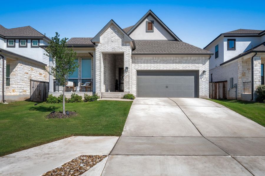 Contemporary residence featuring a light-colored brick and stucco exterior, a dark gray shingled roof, an attached garage with a gray door, a concrete driveway, and a front porch with black railings