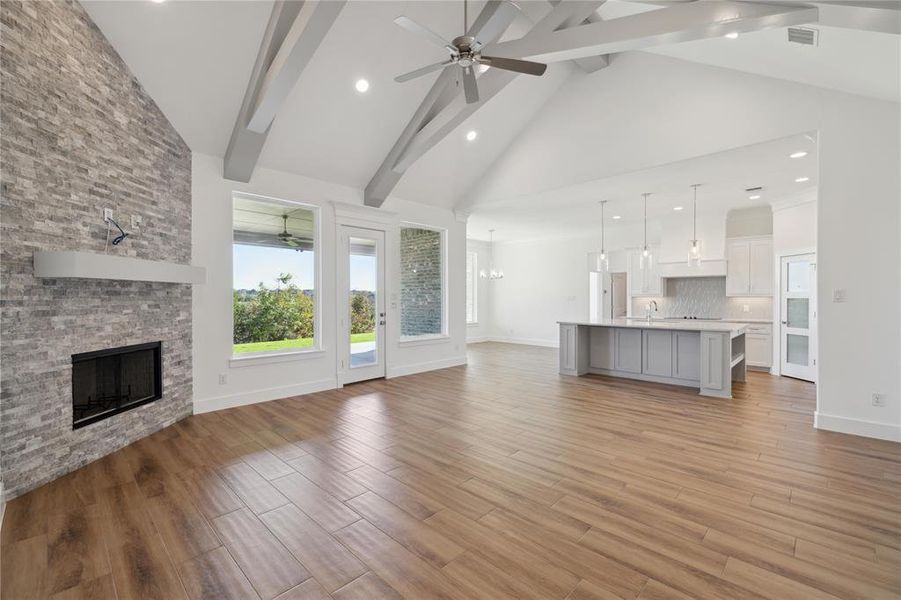 Unfurnished living room featuring light hardwood / wood-style floors, ceiling fan with notable chandelier, beam ceiling, high vaulted ceiling, and a stone fireplace