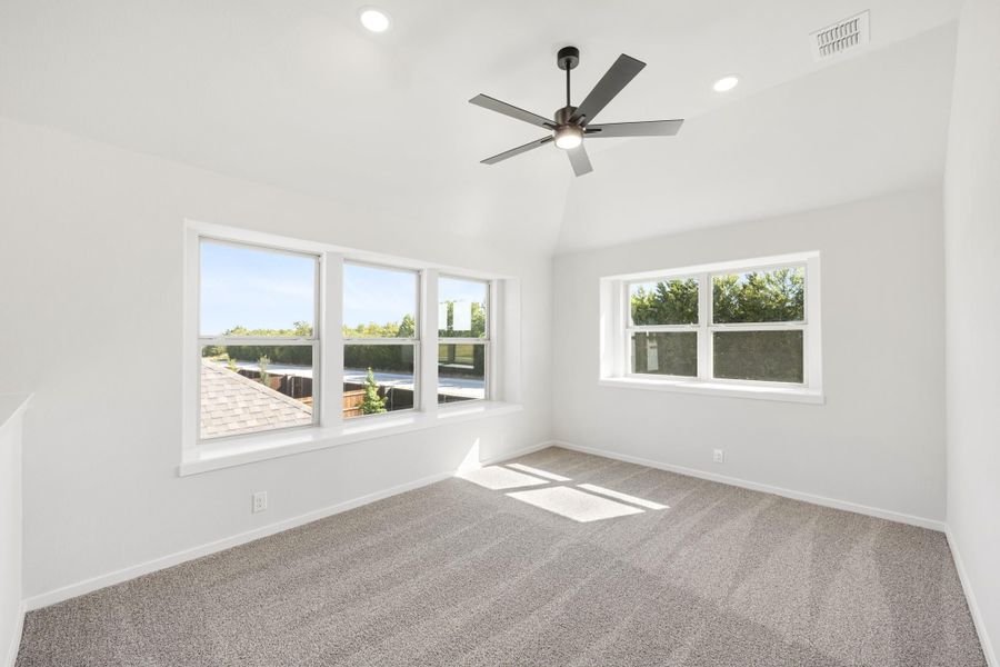 Empty bedroom with vaulted ceiling, ceiling fan, carpet flooring, and multiple windows with natural light
