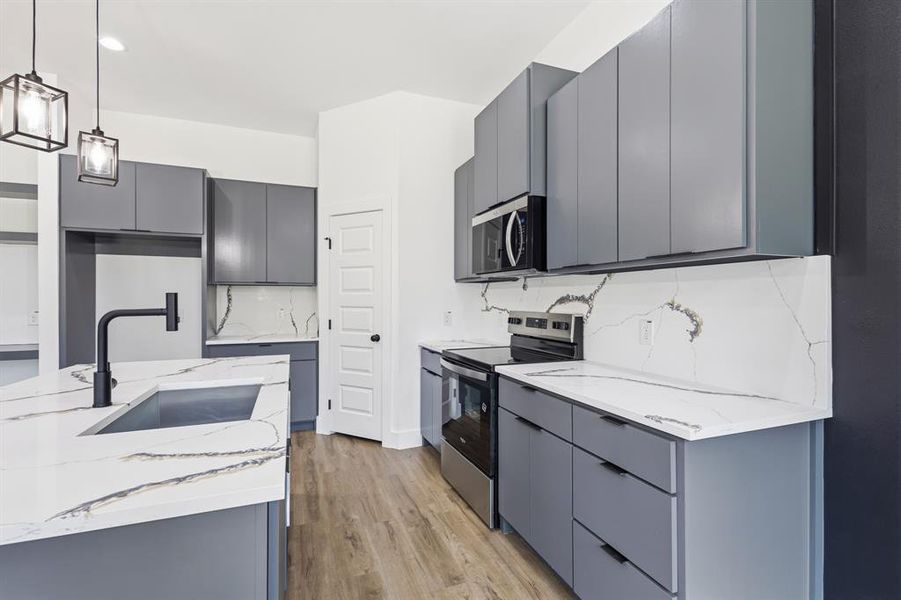Kitchen featuring an island with an undermount sink and matte black faucet, gray cabinetry, stainless steel appliances, white countertops with prominent veining, and wood-finish flooring Kitchen featuring an island with an undermount sink and matte black faucet, gray cabinetry, stainless steel appliances, white countertops with prominent veining, and wood-finish flooring