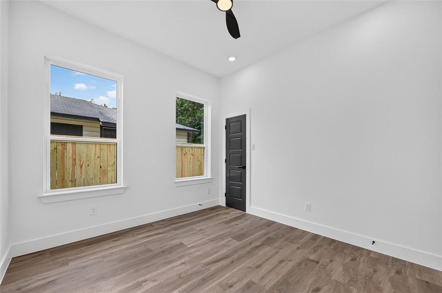 Spacious room featuring wood-finish flooring, two large windows, a ceiling fan, recessed lighting, and a modern dark panel door Spacious room featuring wood-finish flooring, two large windows, a ceiling fan, recessed lighting, and a modern dark panel door