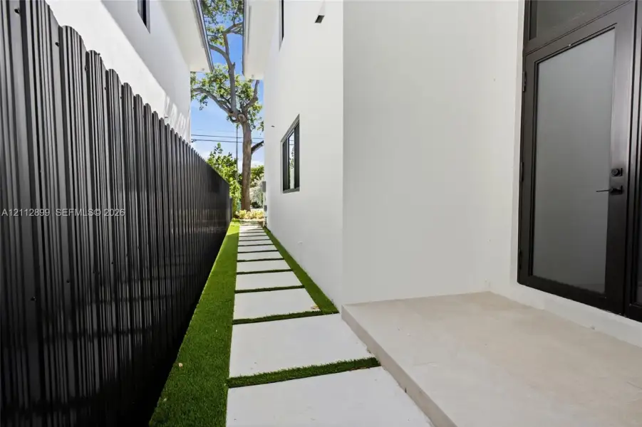 Side exterior featuring white stucco siding, a concrete paver walkway set in artificial turf, a black metal corrugated privacy fence, and a glass-panel entry door
