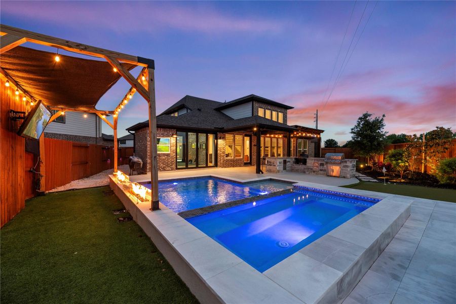 A luxurious pool and hot tub, accompanied by a covered seating area with string lights. The outdoor space also features a built-in grill and lush landscaping, all beautifully illuminated.