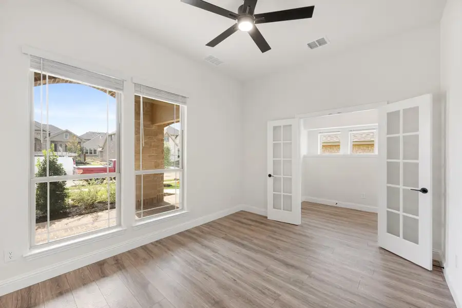 Empty room with hardwood floors, ceiling fan, large windows, and white French doors leading to an adjacent space.