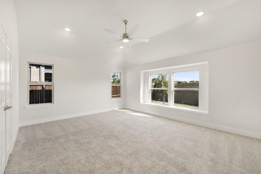 Spacious empty bedroom with vaulted ceiling, ceiling fan, carpet flooring, and multiple windows Spacious empty bedroom with vaulted ceiling, ceiling fan, carpet flooring, and multiple windows