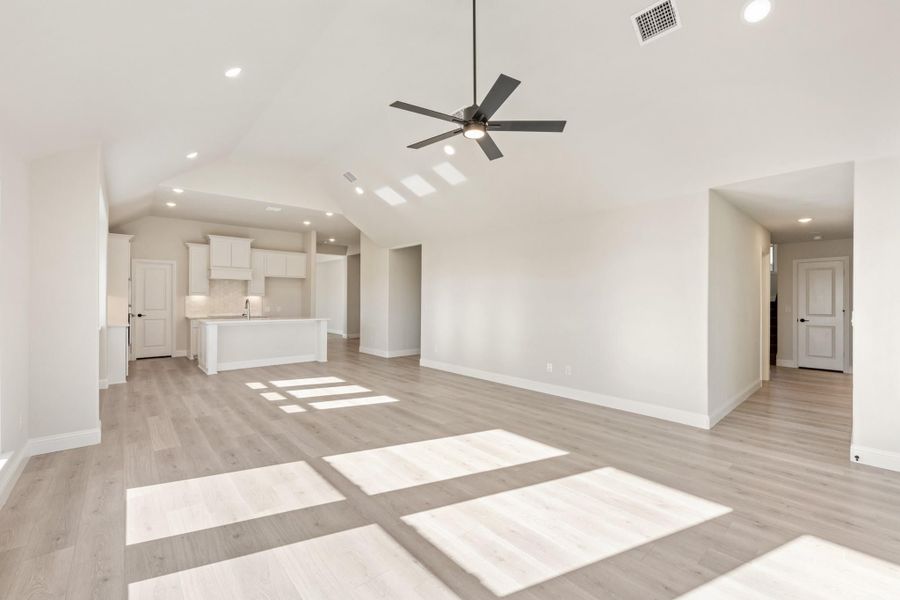 Open living area with vaulted ceiling, ceiling fan, light wood floors, and white kitchen in background