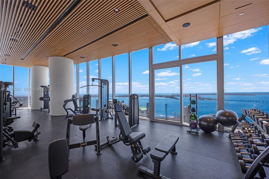 Fitness space featuring floor-to-ceiling windows, slatted wood-finish ceiling, rubberized flooring, and structural columns