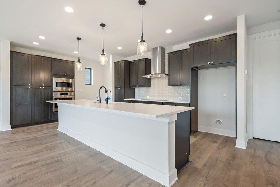 Kitchen featuring hanging light fixtures, wall chimney range hood, stainless steel appliances, dark brown cabinetry, and recessed lighting