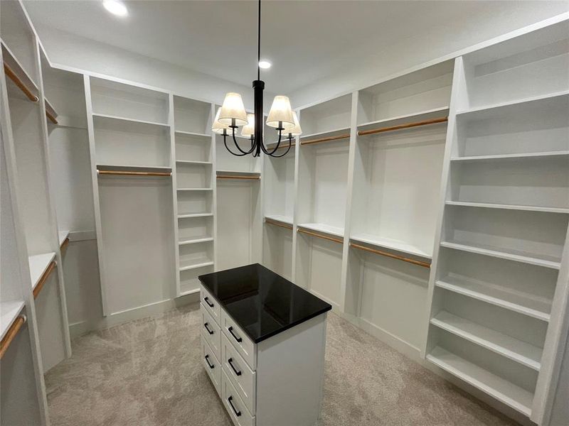 Walk-in closet featuring custom white built-in shelving, hanging rods, an island with a dark countertop and drawers, recessed lighting, and a central chandelier