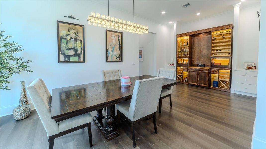 This elegant dining room features a dark wooden table with upholstered chairs, modern artwork on the walls, and a stylish chandelier. It includes a built-in wine storage and display area with warm lighting, set against sleek hardwood floors.