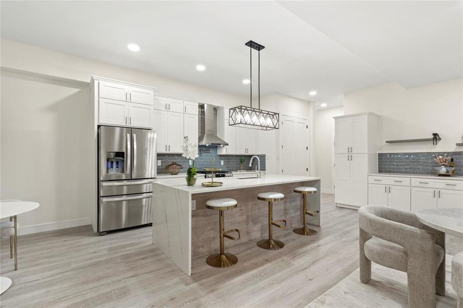 Kitchen with stainless steel fridge with ice dispenser, wall chimney exhaust hood, backsplash, light wood-style floors, and white cabinetry Kitchen with stainless steel fridge with ice dispenser, wall chimney exhaust hood, backsplash, light wood-style floors, and white cabinetry