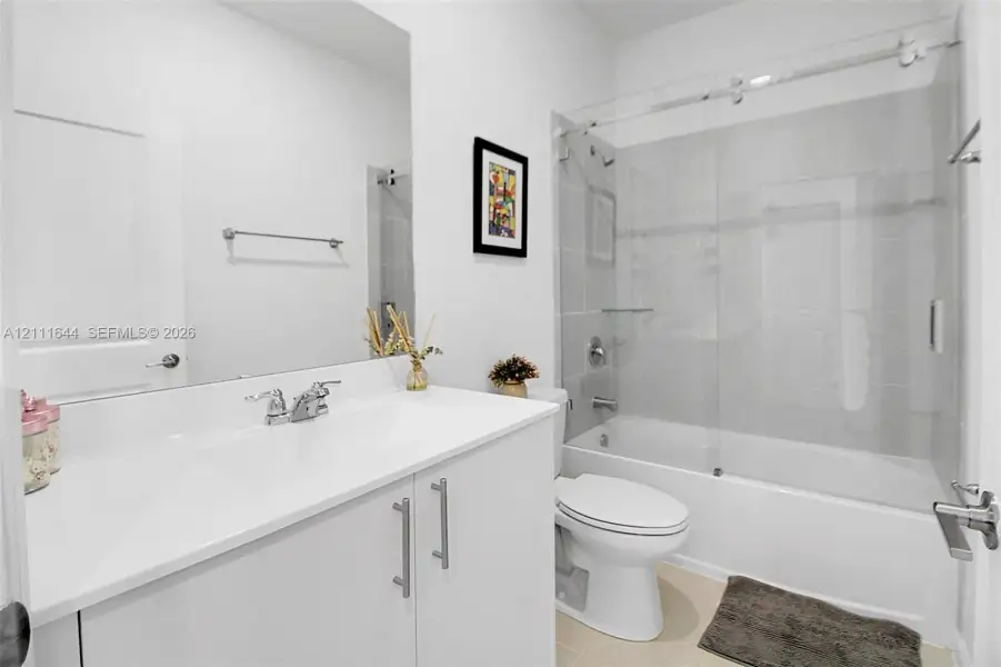 Bathroom featuring a white vanity with a composite countertop, an expansive frameless mirror, and a tub-shower combination enclosed by a sliding glass door with polished chrome hardware Bathroom featuring a white vanity with a composite countertop, an expansive frameless mirror, and a tub-shower combination enclosed by a sliding glass door with polished chrome hardware