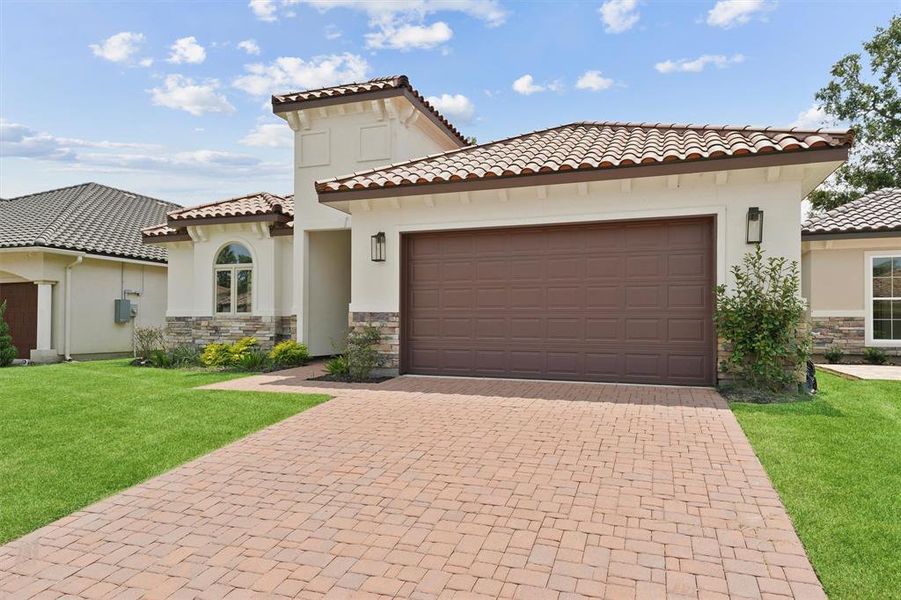 This is a modern single-story home featuring a tile roof, a two-car garage, and a neatly manicured lawn. The architecture includes a stucco finish with stone accents around the windows.