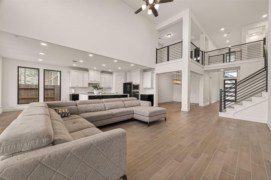 This spacious, open-concept living area features high ceilings, modern finishes, and a sleek kitchen with an island. A large sectional sofa offers ample seating, while the second-floor balcony and staircase add architectural interest. Natural light pours in through large windows, highlighting the elegant flooring.