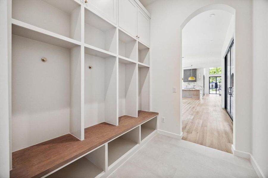 This mudroom is designed for practicality and style, featuring built-in cubbies with storage above and below for shoes, bags, and more. A warm wood bench offers a convenient spot to sit while getting ready, and the crisp white cabinetry adds a clean touch. The arched entryway leads into the open living area, seamlessly integrating the mudroom into the home's overall flow.