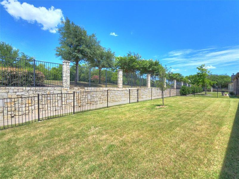 Expansive backyard featuring a stone retaining wall, wrought iron fencing, a verdant lawn, and mature trees