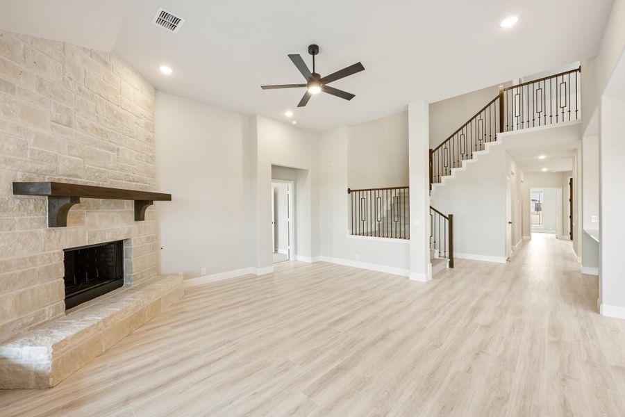 Open living room with stone fireplace, light wood floors, ceiling fan, and staircase with iron railings Open living room with stone fireplace, light wood floors, ceiling fan, and staircase with iron railings