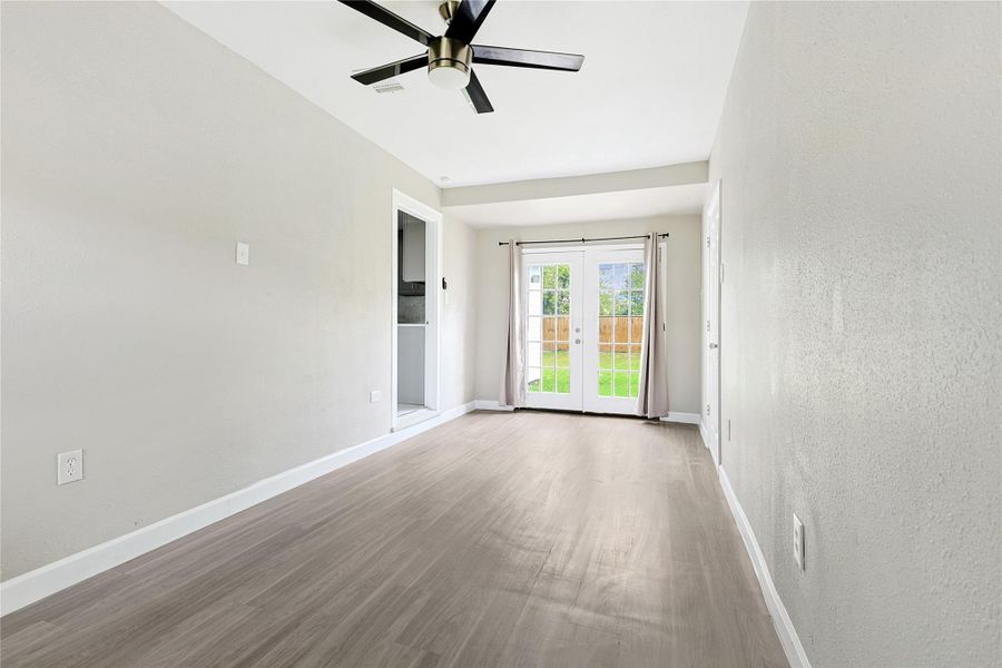 This room features light gray walls and wood flooring, with a modern ceiling fan. It has a bright, open feel thanks to large French doors leading to a backyard, and an adjacent area that could serve as a small dining or reading nook.