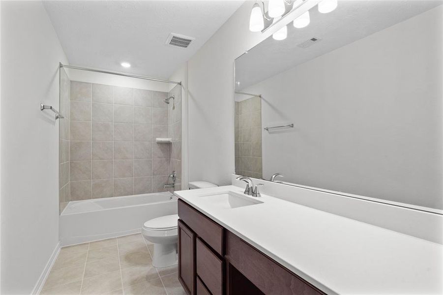 Secondary bath features tile flooring, bath/shower combo with tile surround, stained wood cabinets, beautiful light countertops, mirror, sleek and modern finishes.