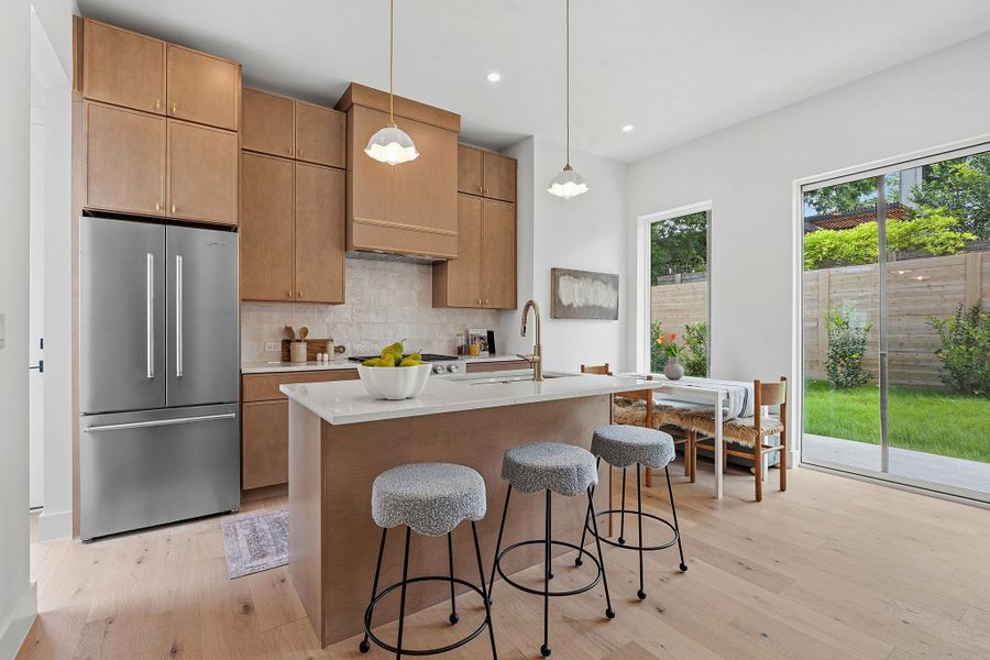 Modern kitchen featuring a large island with an integrated sink, stainless steel appliances, light wood-finish cabinetry, and a full-height tile backsplash