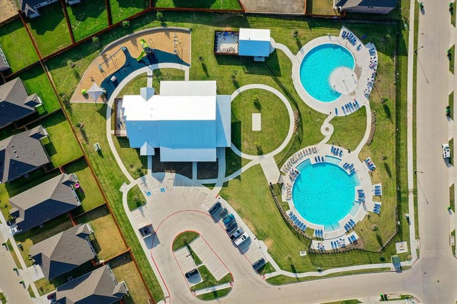 Community recreation area featuring a central building, two swimming pools with surrounding lounge chairs, a playground with shaded structures, and open green spaces