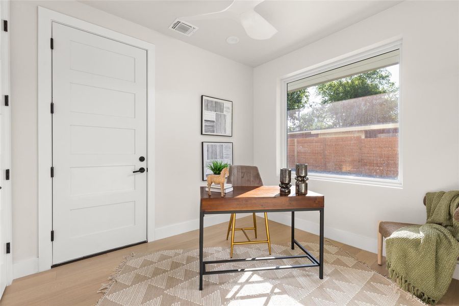 A quiet home workspace features smooth wood flooring and a large window framing backyard views.