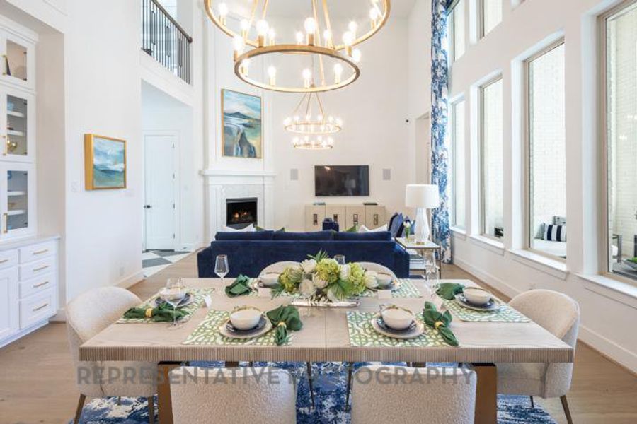 Dining space with an inviting chandelier, baseboards, a towering ceiling, light wood-style floors, and a warm lit fireplace Dining space with an inviting chandelier, baseboards, a towering ceiling, light wood-style floors, and a warm lit fireplace