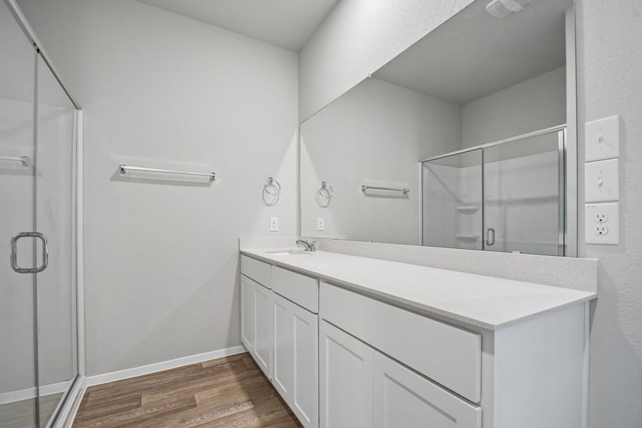 Image of home primary bathroom with wood-look flooring, long white countertop with white cabinets and mirror across the full length. Walk in shower on the left is reflected in the mirror. Towel bar on the wall next to the shower.