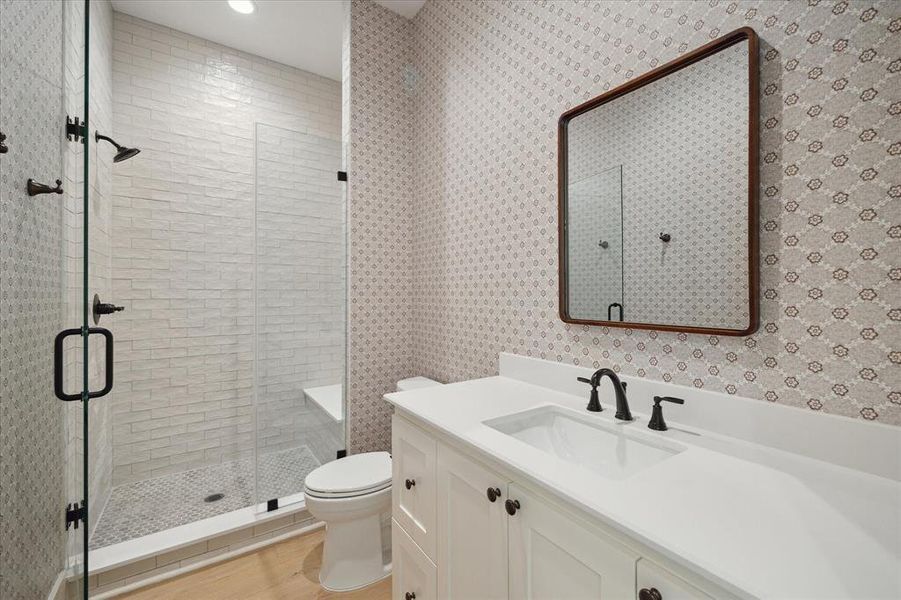 Clean lines and thoughtful design define this first-floor bathroom, which services the adjacent bedroom and main living spaces. The walk-in shower features timeless white subway tile, a built-in bench, and a frameless glass enclosure for a modern and streamlined aesthetic. Patterned wallpaper adds character, while the vanity boasts white cabinetry, a quartz countertop, and matte black fixtures. Warm toned flooring and a custom-framed mirror enhance the cohesive and welcoming feel of the space.