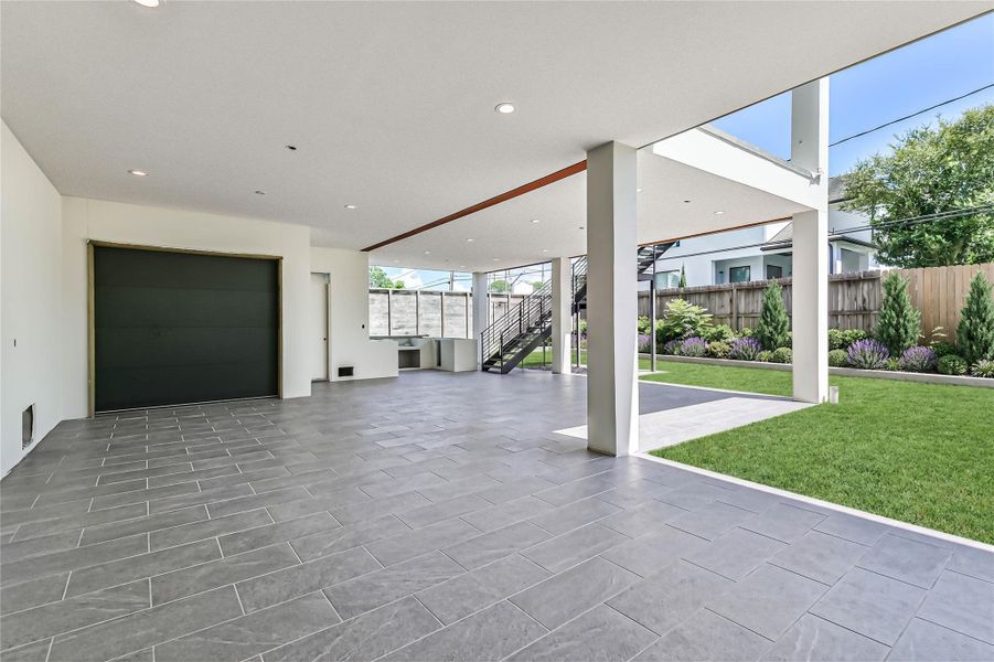 The sweeping covered backyard terrace will feature stained concrete floors, an integrated summer kitchen,  recessed lighting, built-in speakers and access to the storage space under the home. The voluminous shaded footprint looks directly onto a manicured green lawn with vibrant, structured landscaping, providing a private and protected environment built for seamless grand-scale entertaining. This is a progress photo from May 26th with *virtually rendered floors and landscaping*