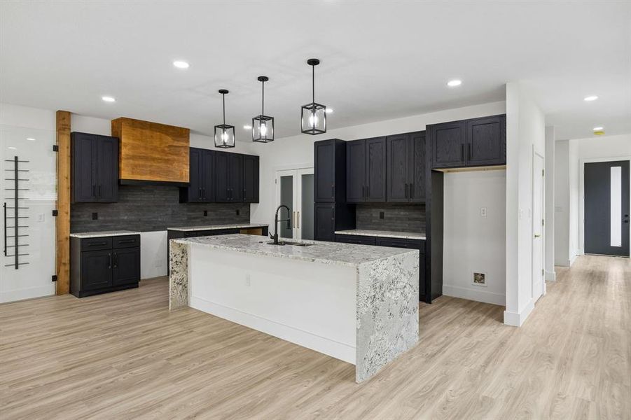 Modern kitchen featuring a large waterfall island with a stone countertop, dark cabinetry, and a light wood-finish range hood Modern kitchen featuring a large waterfall island with a stone countertop, dark cabinetry, and a light wood-finish range hood
