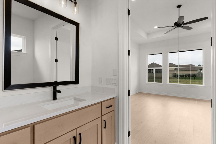 A pristine bathroom vanity with a spacious mirror complements the bright bedroom with a ceiling fan and ample natural light.