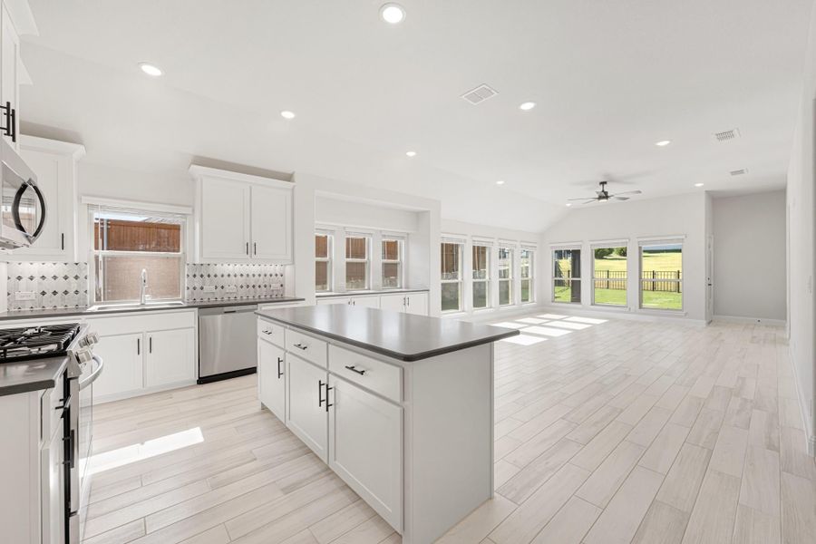 Open kitchen with white cabinets, center island, dark countertops, and light wood floors opening to living area with large windows