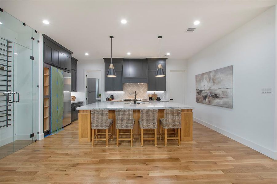 Kitchen with tasteful backsplash, custom exhaust hood, a kitchen island with sink, light hardwood / wood-style floors, and hanging light fixtures