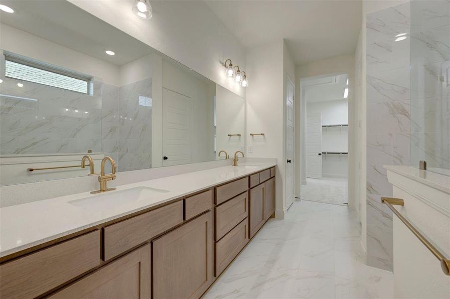 Double vanity with light wood-finish cabinetry, white countertops, and gold-tone fixtures Double vanity with light wood-finish cabinetry, white countertops, and gold-tone fixtures