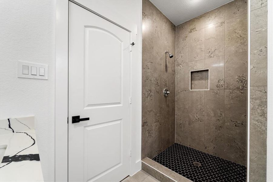 This modern bathroom features a sleek walk-in shower with floor-to-ceiling marbled tile and bold black hexagonal flooring. Matte black hardware and a crisp white door complete the stylish and functional space.