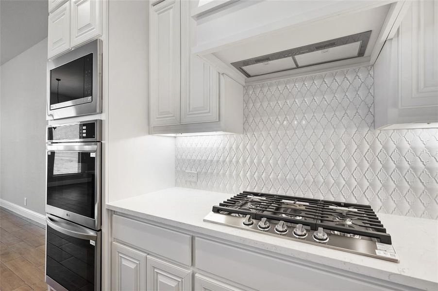 Kitchen with range hood, stainless steel appliances, white cabinetry, light wood finished floors, and light stone counters