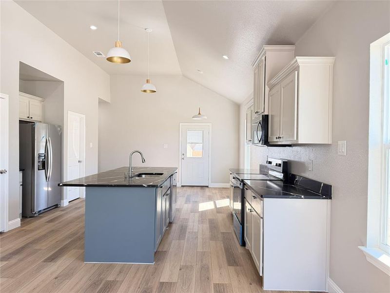 Kitchen featuring stainless steel appliances, a center island with sink, light wood finished floors, decorative light fixtures, and vaulted ceiling
