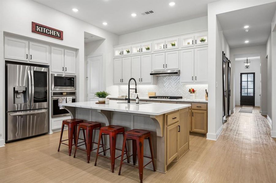 Open-concept kitchen featuring an oversized island with countertop seating, stainless steel appliances, white shaker cabinetry with under-cabinet lighting, and wood-finish flooring Open-concept kitchen featuring an oversized island with countertop seating, stainless steel appliances, white shaker cabinetry with under-cabinet lighting, and wood-finish flooring