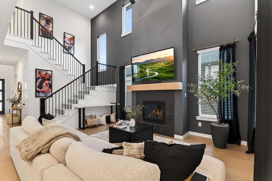 Living room featuring a tiled fireplace, a towering ceiling, light wood-style flooring, stairs, and healthy amount of natural light Living room featuring a tiled fireplace, a towering ceiling, light wood-style flooring, stairs, and healthy amount of natural light