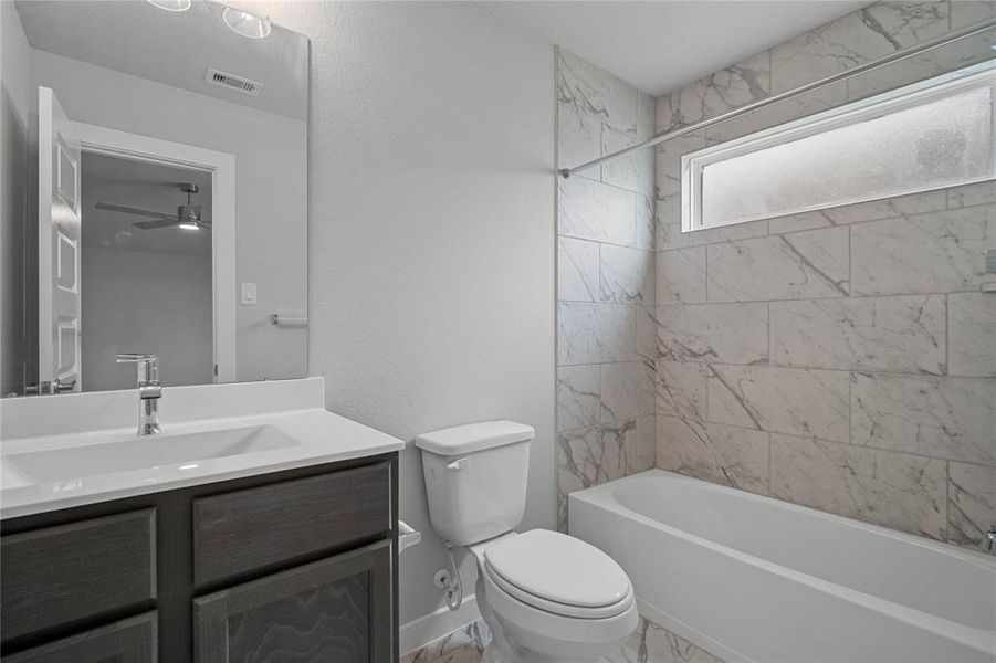 Secondary bath features tile flooring, bath/shower combo with tile surround, stained wood cabinets, beautiful light countertops, mirror, dark, sleek fixtures and modern finishes.