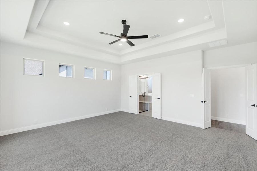Spacious bedroom with tray ceiling, ceiling fan, gray carpet, and ensuite bathroom doorway