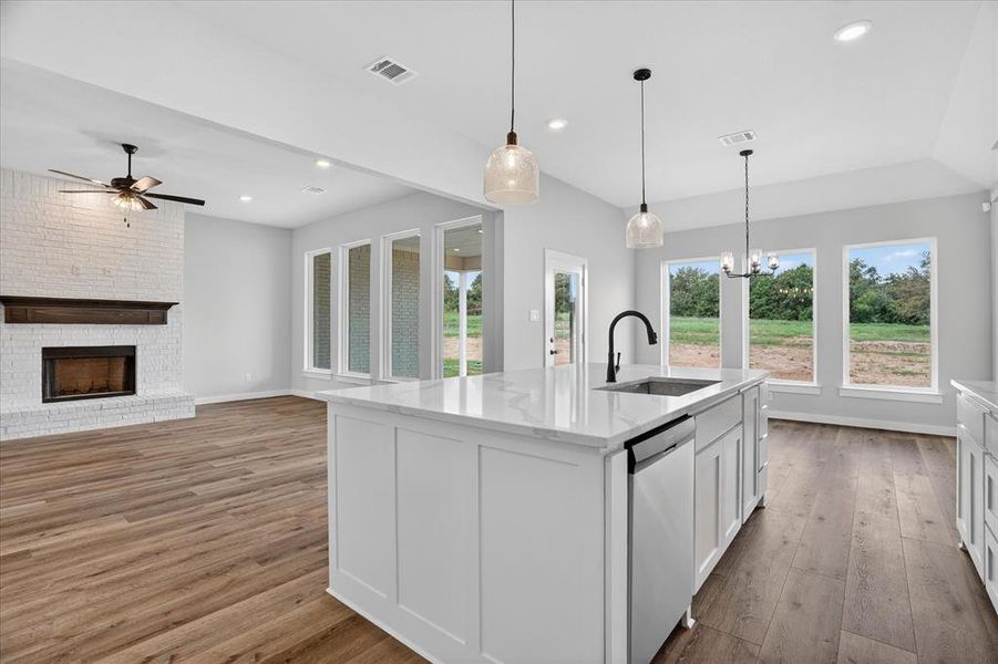 Spacious open concept living area featuring a kitchen island with sink, white shaker cabinetry, stainless steel appliances, wood-finish flooring, and a whitewashed brick fireplace