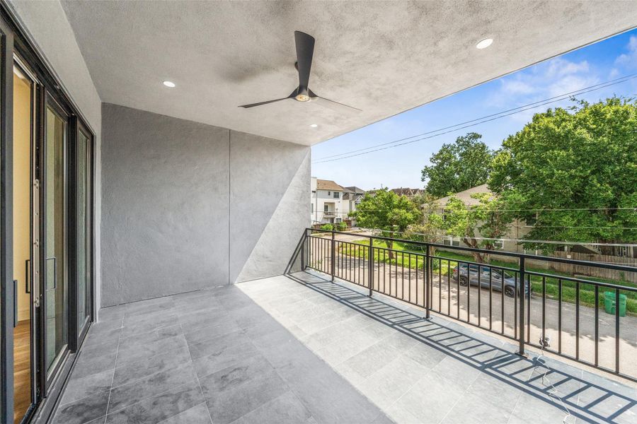 Balcony with sleek tile flooring and a ceiling fan. It features a metal railing and offers a pleasant view of the neighborhood, with trees and houses visible in the background. Perfect for outdoor relaxation.