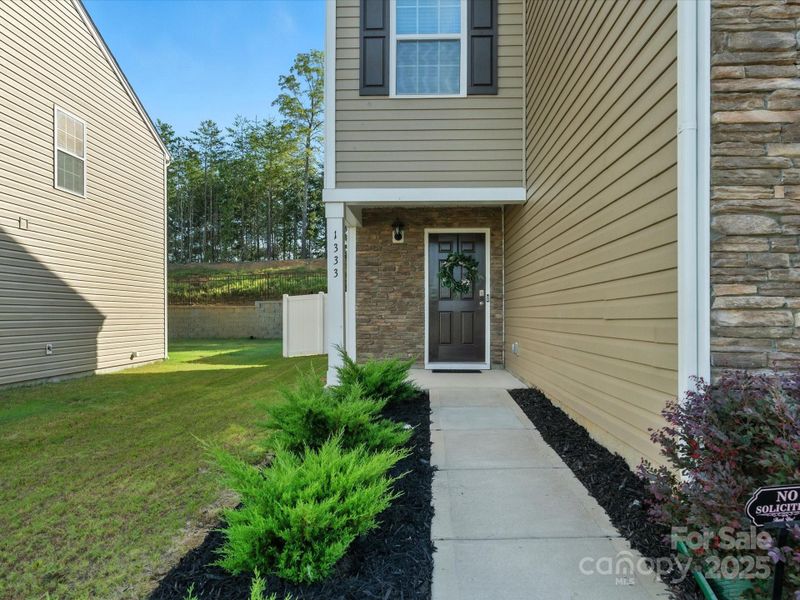 Welcoming covered entry features stone surround and professional landscaping with evergreen plantings. Clean walkway and attractive front door with seasonal wreath create an inviting first impression.