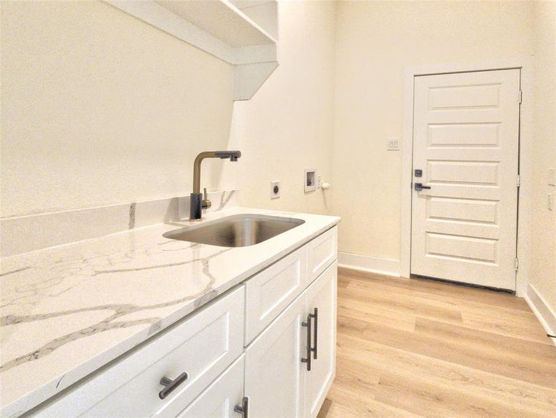 Utility room featuring light wood-finish flooring, white paneled cabinetry with dark hardware, a stainless steel sink, a modern dark faucet, and a light-toned countertop with veining