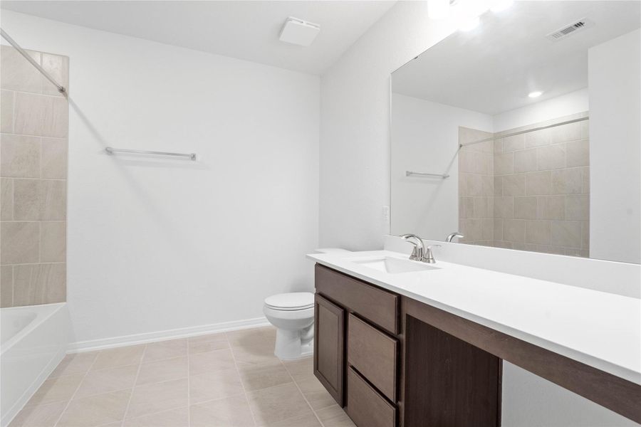 The secondary bathroom offers tile flooring, a bath/shower combination with a stylish tile surround, rich stained wood cabinetry, elegant light countertops, a mirror, and sleek fixtures with modern finishes.