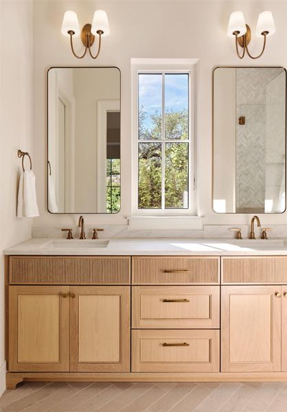 Primary bath features dual vanity, quartz counter, and custom cabinetry with designer hardware. Cabinets feature reeded panels and feet. Designer mirrors illuminated by lamp shade style glass sconces.