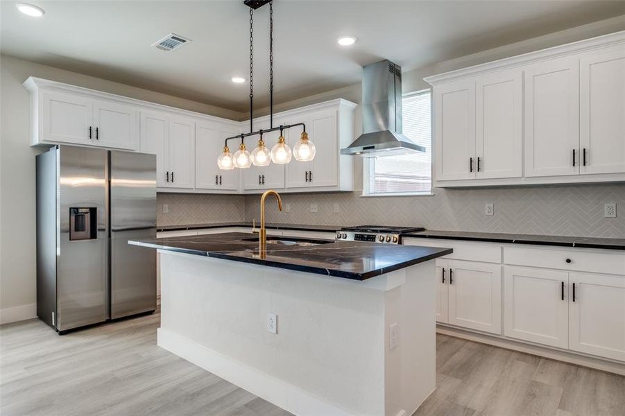 Kitchen featuring stainless steel appliances, decorative light fixtures, a center island with sink, wall chimney exhaust hood, and white cabinetry